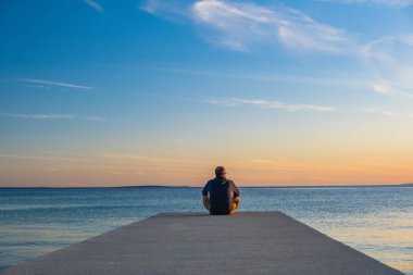 Adam rıhtımın kenarında oturmuş meditasyon yapıyor. Gün batımında deniz ufku, Pag Adası, Adriyatik denizi, Hırvatistan.