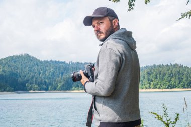 DSLR kameralarıyla gölde dolaşan kapüşonlu yetişkin bir adam. Çekimler, güzel bir yaz günü, yarı vücut portresi, manzara fotoğrafı konsepti.