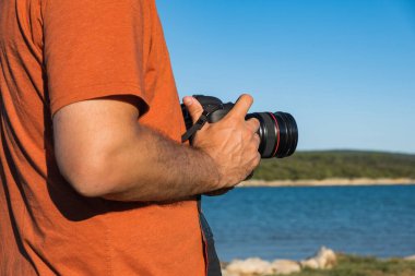 DSLR kamerasını elinde tutan isimsiz bir adam deniz kıyısının yanında, yüzü yok, vücut detayı yok, manzara fotoğrafçılığı konsepti