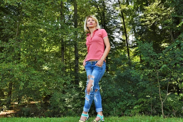 穿着破旧牛仔裤和彩色凉鞋的年轻苗条女子站在树林里 — 图库照片