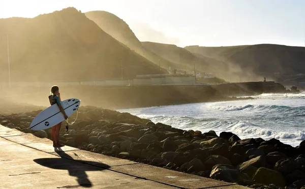 2018年 西班牙大加那利岛的拉斯帕尔马斯 一个拿着冲浪板的女孩站在石头附近的日日里的海洋附近 — 图库照片
