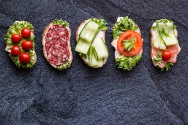 Sosis, peynir, pastırma, domates, marul, koyu zemin üzerinde salatalık ile taze sandviç. Yatay yönlendirme