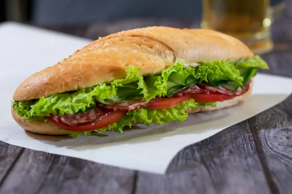 Sanduíche submarino fresco com salsicha, queijo, bacon, tomate, alface, pepino e cebola em um fundo de madeira escura. Caneca de cerveja — Fotografia de Stock