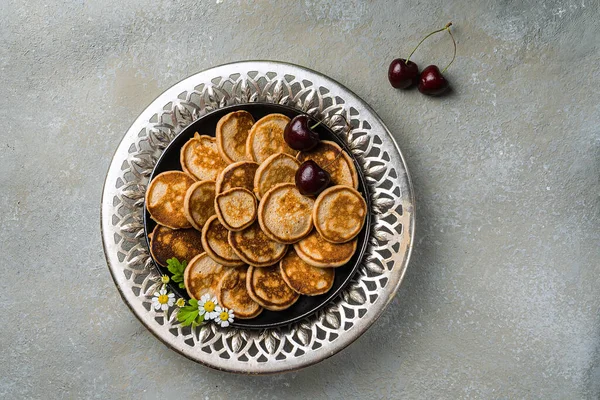 Um café da manhã caseiro na moda com pequenas panquecas close-up. Mini panquecas, cerejas, chá na mesa em uma placa de ornamento com aberturas. Orientação horizontal — Fotografia de Stock