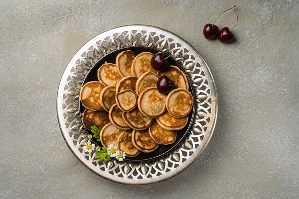 Um café da manhã caseiro na moda com pequenas panquecas close-up. Mini panquecas, cerejas, chá na mesa em uma placa de ornamento com aberturas — Fotografia de Stock