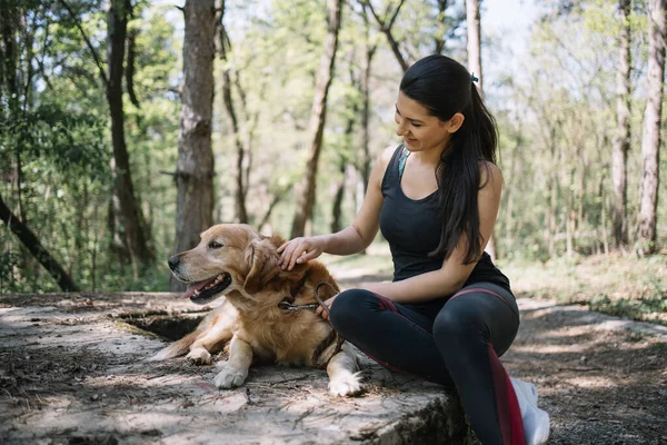Kız ormanda otururken köpeğini okşuyor.