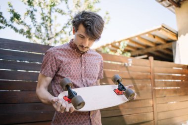 Esmer adam kaykayı tutuyor ve tekerlekleri kontrol ediyor.