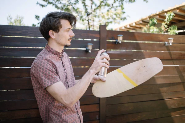Mladý muž sprejování barva přes dřevěný skateboard — Stock fotografie