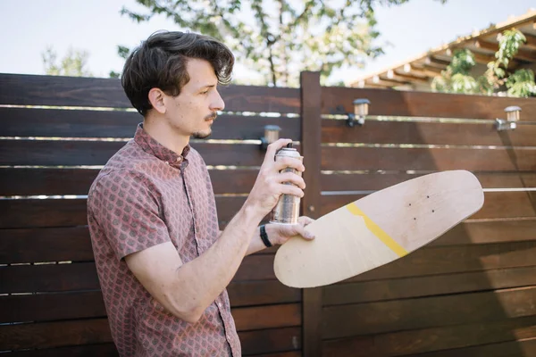 Mladý muž sprejování barva přes dřevěný skateboard — Stock fotografie