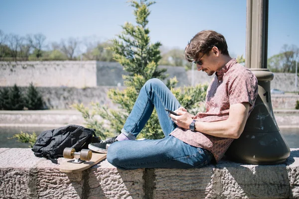 Oldalnézet az ember ül a kő ül és nézi a telefont — Stock Fotó