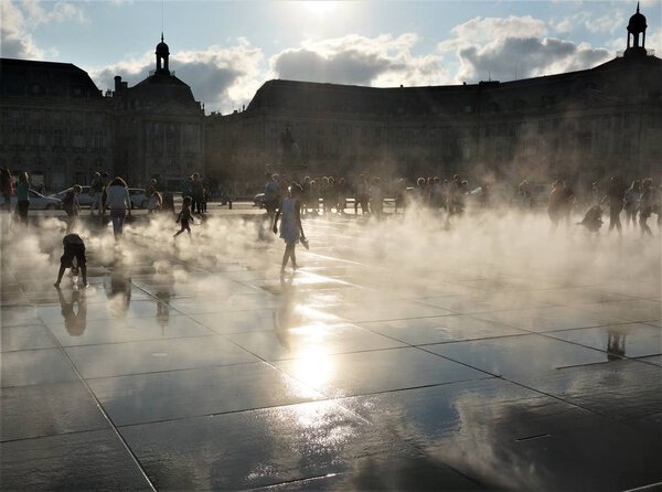 silhouettes of people and buildings in the fog and a reflection of the sky in the fountain in Bordeaux on a hot day