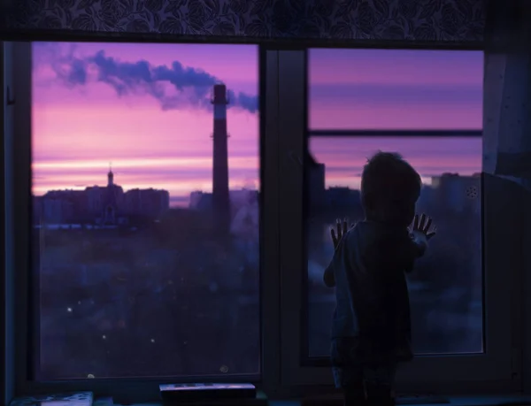 Una silueta de un niño (niño pequeño) en la ventana mira el amanecer rosa y ve humo y casas urbanas . — Foto de Stock
