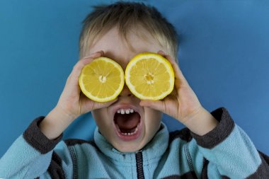 Kafkas çocuk Toddler sarışın mavi arka plan üzerinde gözleri dilimlenmiş limon ile komik yüz yapma. Duygu kavramı, sağlık, sululuk, narenciye vitamintazeliği
