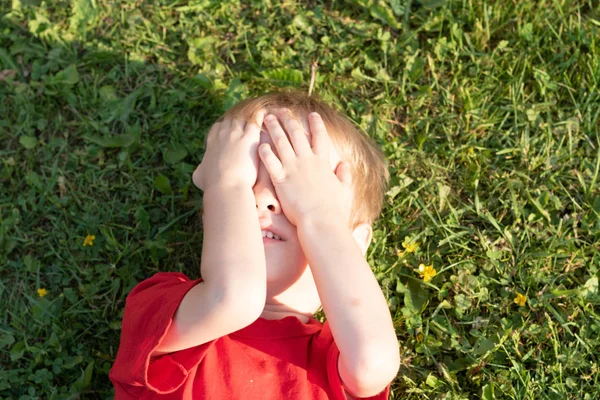 Europeu loiro menino fechado seus olhos com as mãos deitadas na grama — Fotografia de Stock