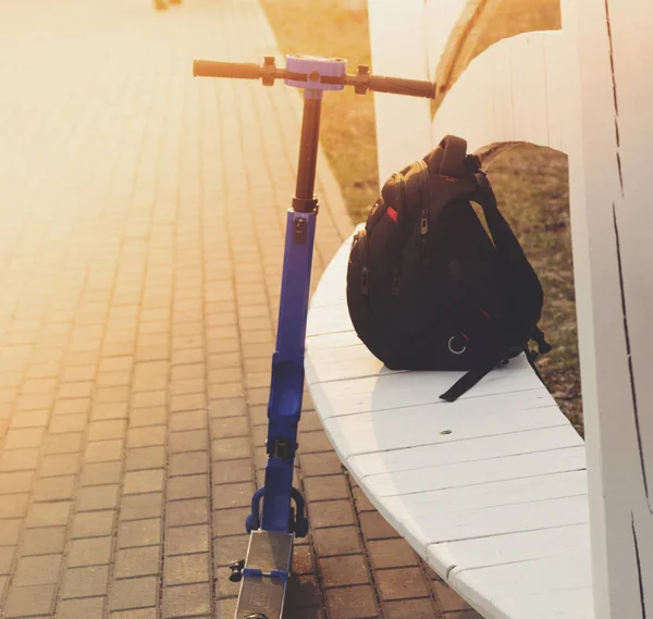 An eco-friendly electric blue scooter stands in a park leaning on a bench, on which lies a black urban backpack. Evening, sunset. Vertical. No people no body