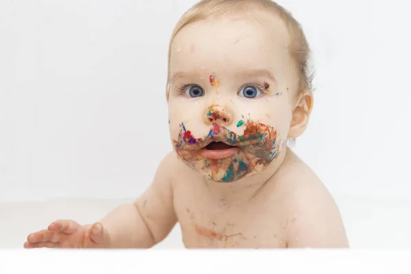 La cara del niño está manchada con pintura multicolor, que el niño comió y pintó sobre un fondo blanco. El concepto de creatividad y brillo, desarrollo desde el nacimiento . — Foto de Stock