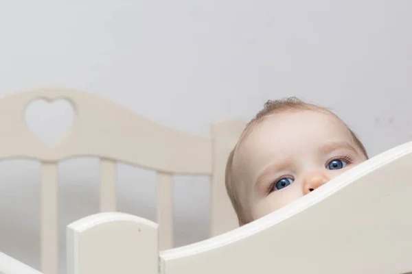The face of a Caucasian baby toddler peeps out from behind the crib on a gray background. Blue eyes are looking at the camera. — Stock Photo, Image