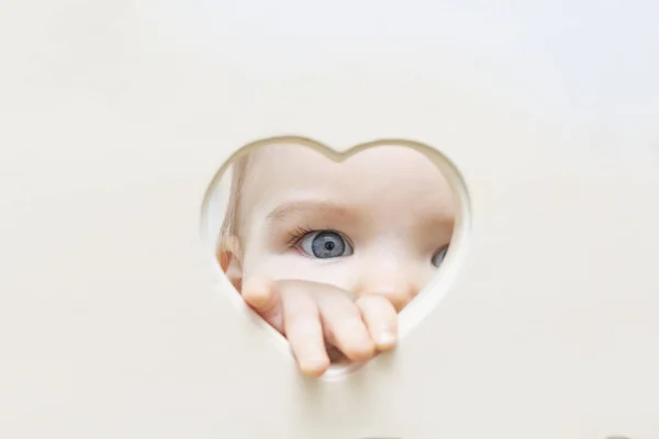 아기의 얼굴, 눈과 손은 중앙에 있는 하트 모양구멍에서 들여다보습니다. 한 어린이가 심장 구멍을 통해 카메라를 들여다봅니다. 사랑과 건강 개념, 모형, 포스터, — 스톡 사진