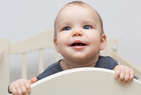 Retrato europeu caucasiano feliz sorrindo bebê criança com olhos azuis em um fundo branco olhando acima da câmera segurando para berço cama. Idade - 10 meses - 1 ano — Fotografia de Stock