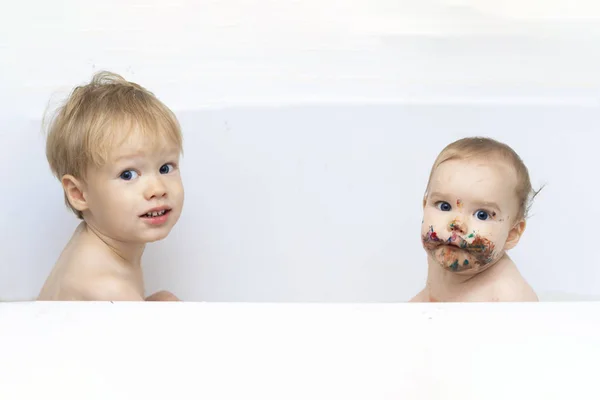 Δύο παιδιά, αδέρφια κάθονται στο μπάνιο, το ένα είναι καθαρό, το άλλο βρώμικο. Το πρόσωπο του παιδιού είναι χρωματισμένο με πολύχρωμη μπογιά, την οποία έφαγε το παιδί. Σε λευκό φόντο — Φωτογραφία Αρχείου