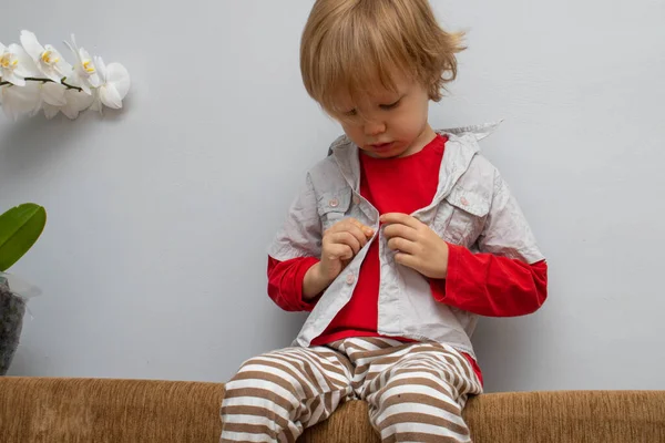 Concepto de independencia de la infancia, niño pequeño abotonándose en la camisa, sujetando sus botones, sobre fondo gris blanco. El niño se pone su propia ropa. — Foto de Stock