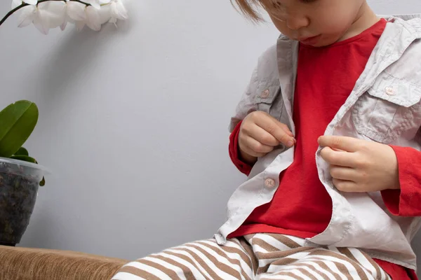 Concepto de independencia de la infancia, niño pequeño abotonándose en la camisa, sujetando sus botones, sobre fondo gris blanco. El niño se pone su propia ropa. — Foto de Stock