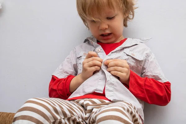 Concepto de independencia de la infancia, niño pequeño abotonándose en la camisa, sujetando sus botones, sobre fondo gris blanco. El niño se pone su propia ropa.. — Foto de Stock