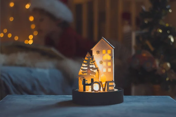 lamp in form of a house with text home on background of a cozy apartment interior with a blurred bokeh garland. Blurred child reads a book. The dark trendy style is toned. Home and family concept