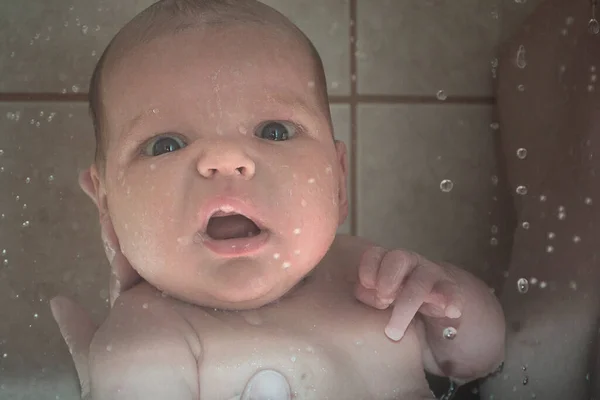 The first bathing of the newborn with a shower. Emotional facial expression of a baby under drops of water. Fear of water and bathing