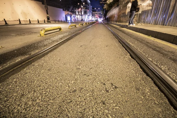 Villamos sínek aszfalton. Sétáló emberek és közlekedési lámpák a járdán. Éjszaka hosszú expozícióval filmezve. — Stock Fotó