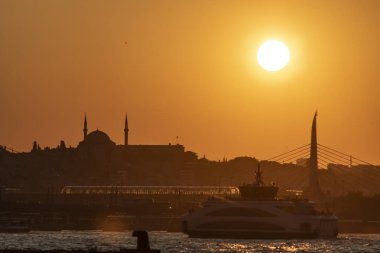 İstanbul Denizi üzerinde gün batımında metro köprüsü ve buharlı gemi görüntüsü. Güneş arka planda parlıyor..