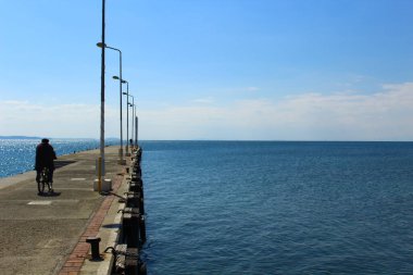 Deniz kenarı ve beton iskele. Geleneksel giyimli adam iskelede bisiklet sürüyor. Açık havada güneşli bir fotoğraf..