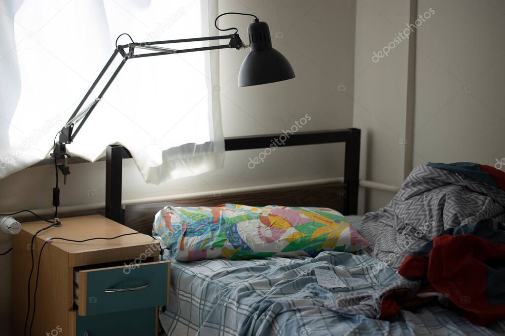 Untidy single bedroom. Alone single man. There is a table lamp by the window. Photographed during the day.