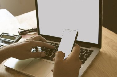 bir adamın eli cep telefonu tutuyor, diğer taraftan dizüstü bilgisayarın klavyesi üzerinde, masada bir hesap makinesi ve gözlük, monitör ve cep telefonu metin için bir yer var