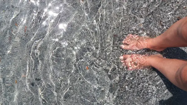 Kadın ayak banyosu yapıyor, ayakları şeffaf suyla temiz bir sahilde sırılsıklam oluyor. 