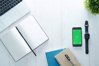 Not defteri, akıllı telefon, saat, fare, klavye ve malzemelerle dolu ofis masası, beyaz ahşap masaya izole edilmiş. Kopyalama alanı olan üst görünüm, beyaz, temiz, düz uzanma.