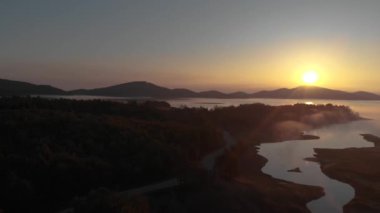 Etrafında tepeler ve asfalt yollar olan gölün üzerinde güzel bir gün doğumu manzarası var..