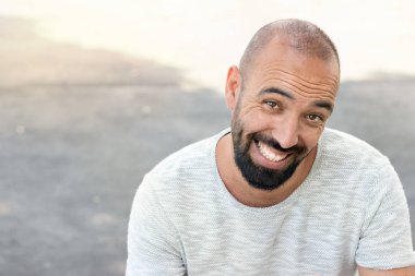 Sakallı ve ultra kısa vızıltılı bir adamın portresi, komik bir şekilde gülümseyen, kamera görünümlü, bir t-shirt giyen, açık havada.