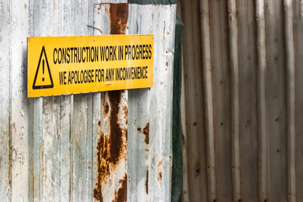 A construction work in progress sign board over a corrugated wall panel, outdoors.