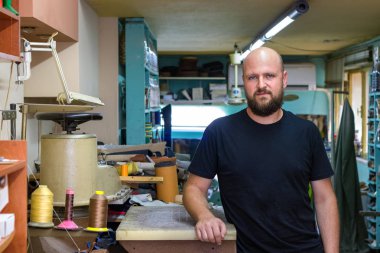 Sakallı, atölyesinde ayakta poz veren, ayakkabıcı kel bir adamın portresi.