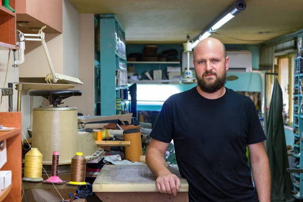 Sakallı, atölyesinde ayakta poz veren, ayakkabıcı kel bir adamın portresi.