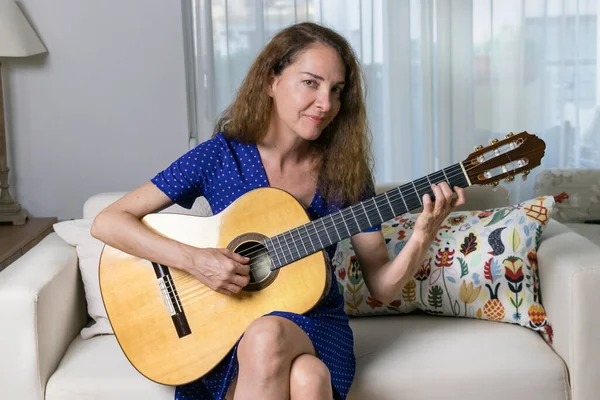 Female guitar teacher playing an acoustic guitar at home, looking camera