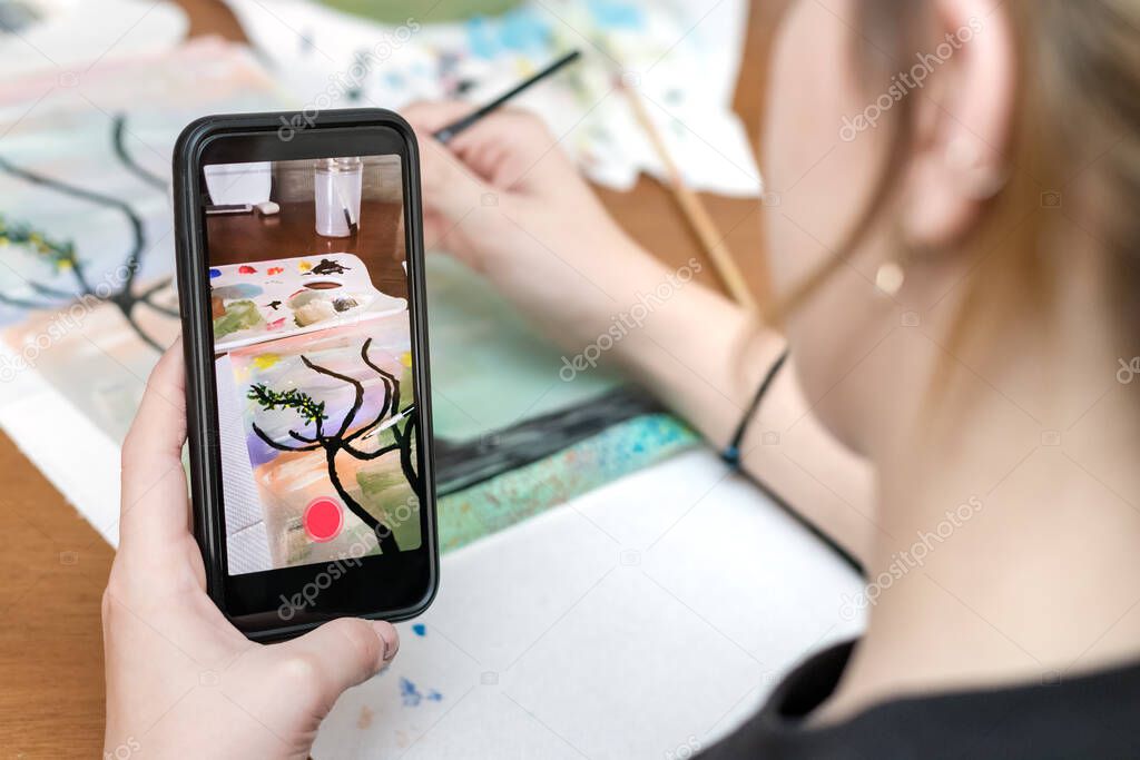 Teenage female hand holding a smart phone taking photos - video of herself drawing a paint. 