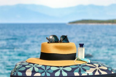 Maske, el dezenfektanı ve plaj arka planında güneş gözlüğü olan bir çantada hasır şapka..