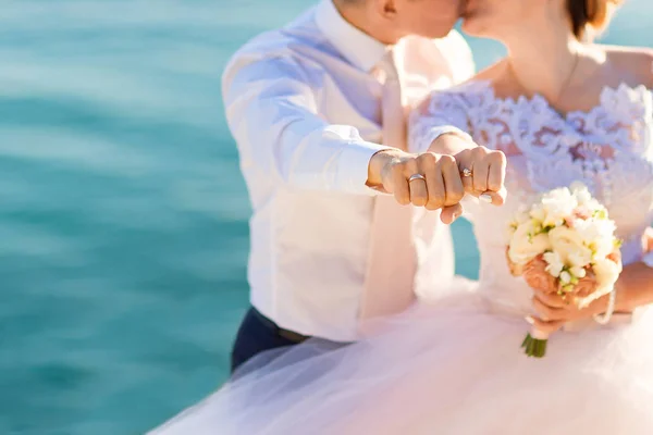 Kissing newlyweds reach out their fists with wedding rings on the the sea background — ストック写真