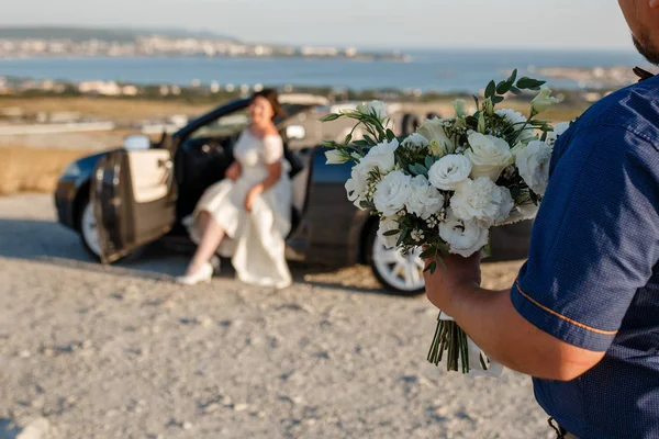 Groom holding wedding bouquet flowers and see to bridge sitting in wedding car — Stock Photo, Image