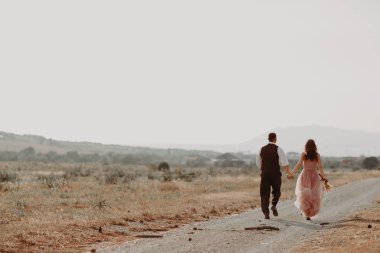 Gelin ve damat el ele tutuşup gelinliklerle koşuyorlar. Gelin elinde bir buket çiçek tutuyor.