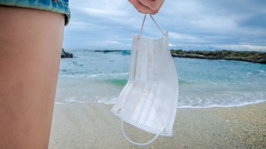 Tıbbi maske tutan ve kumsalda dinlenen bir kadın. Genç bir kadın koruyucu maskeyle ufka bakıyor. COVID-19 için alarm durumu. Tatil ve Coronavirüs durum konseptini çöz