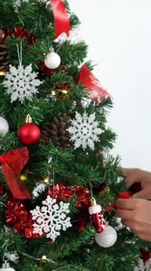 Dikey Asyalı kadın oturma odasındaki süslerle Noel ağacını süslüyor. Mutlu Noeller ve mutlu yıllar konsepti. Kız ladin dallarına Noel süsleri takıyor. Aile sıcacık anları