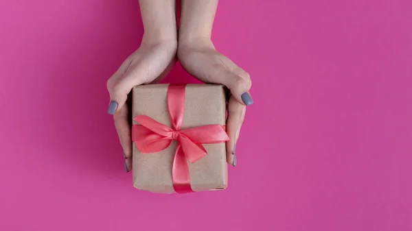 Menina segurando um presente em mãos, mulheres com caixa de presente em mãos envolvidas em papel artesanal decorativo com um laço de fita vermelha amarrada em fundo de papelão colorido, vista superior, férias conceito, amor e cuidado — Fotografia de Stock